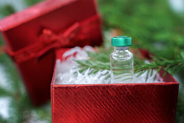 Best gift for Christmas. Vial with new tested vaccine for covid-19 coronavirus, dangerous flu, infection. Small red holiday box with bow. Green branches of new year tree, decorations. White plaid © velirina