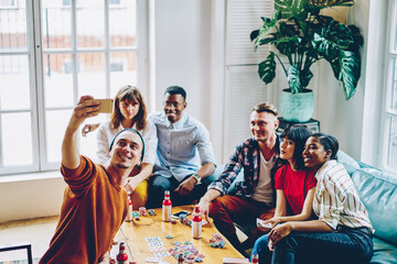 Fototapeta na wymiar Group of diverse players looking at camera while photographing themself during weekend poker gaming, youthful hipsters enjoying leisure drinks meeting in cozy interior shooting vlog party video