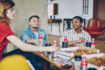 Fototapeta premium Hipster friends sitting at table with delivered pizza and alcohol drinks discussing board game with chips for playing poker during weekend, multicultural people enjoying gaming entertainment