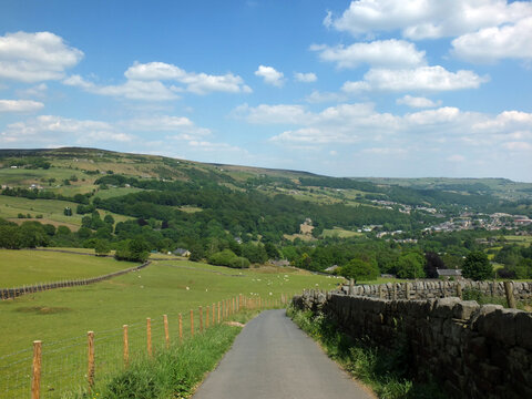 country lane running downhill surrounded by fields with sheep with a view of the town of mytholmroyd surrounded by woods and fields in the calder valley west yorkshire