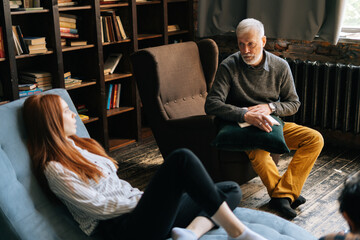 Professional gray-haired man psychologist consulting patient during psychological consultation in cozy dark office room. Bearded mature psychotherapist taking notes and giving advices to patient.