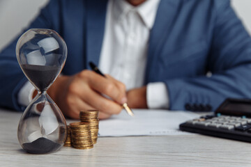 Businessman signing important contract in office. Stack of coins and hourglass on desk close-up....