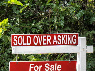 A red real estate for sale and Sold Over Asking sign against a leafy background