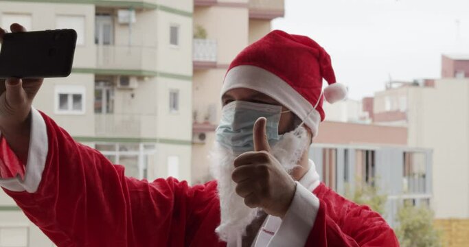 

Man dressed as Santa Claus with medical mask taking a selfie and making the ok symbol with a smartphone Christmas and coronavirus