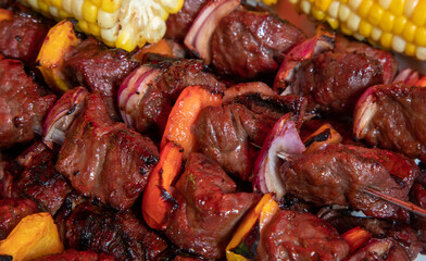 close up of cooked beef tenderloin skewers or shish kebabs with red and yellow peppers and onion pieces ready to serve with cobs of corn