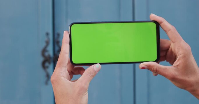 Close up view of female hands holding smartphone with green tamplate screen horizontally. Concept of mockup and chroma key. Blue door at background.