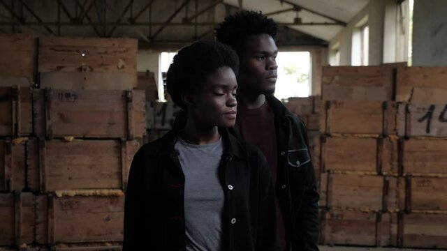 attractive African-American couple wearing black denim jackets poses for camera against wooden boxes in storehouse slow motion