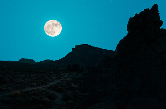 At night in the middle of the crater of the Teide volcano in Spain. There are a lot of rocks. The full moon shines on the backlit landscape.