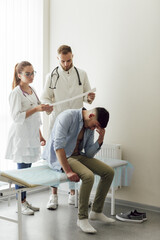 Obraz premium Two doctors, a man and a woman, read a bad cardiogram of a young patient in a hospital office. A young man on examination at the hospital, sitting on a couch with his head in his hands