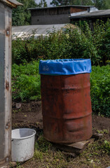 Obraz premium old red rusty barrel with blue bag inside stands in a green garden among greenhouses