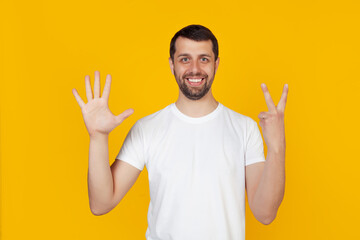 Modern young man with a beard in a white tank top shows number seven with fingers on hand, smiling confidently and happily, looking into the camera. The man shows seven fingers. Number 7