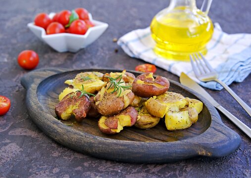 Crash Hot Potatoes, boiled potatoes in a peel, crushed and baked with olive oil and herbs on a wooden plate on a dark concrete background. Australian cuisine. Potato recipes.