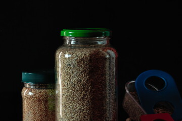 transparent flasks lit from the side, filled with birdseed and millet, next to blue and red eaters for birds, on a black background.