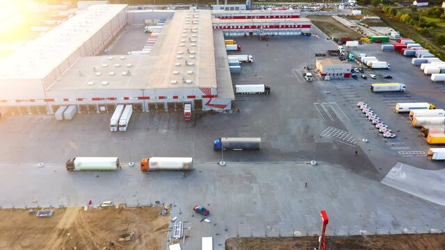 Large logistics park with a warehouse - loading hub. Semi-trucks with freight trailers standing at the ramps for loading/unloading goods at sunset. Aerial hyper lapse (motion - time lapse)