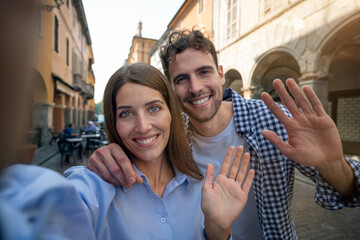 An young happy romantic couple of tourists in love is making selfie or technology video call with smartphone to friends or family while having fun and visiting new places together in old city center.