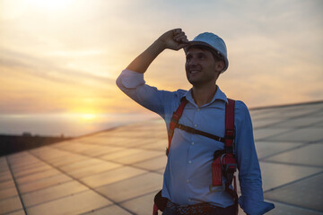 Maintenance assistance technical worker in uniform is smiling satisfied of his work after checking operation and efficiency performance of photovoltaic solar panels on roof at sunset.
