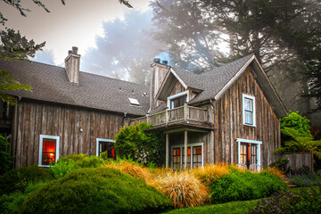 Rustic wood house surrounded by tall trees with fog and smoke and lights shinning out from windows
