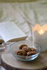 Wooden tray with bowl of chocolate chip cookies and lit candle on a bed. Open book in the background. Selective focus.