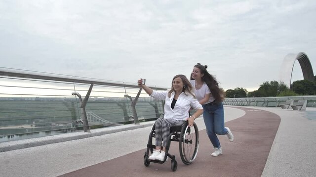 Joyful teenage daughter driving smiling handicapped mother on wheelchair while disabled woman shooting live stream on smartphone. Cheerful females having fun shooting live video on pedestrian bridge
