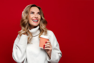 Studio portrait of charming woman in white sweater, wear earmuffs, holding paper coffee cup takeaway, isolated on red background, looking at blank space for advertising. Winter sale.