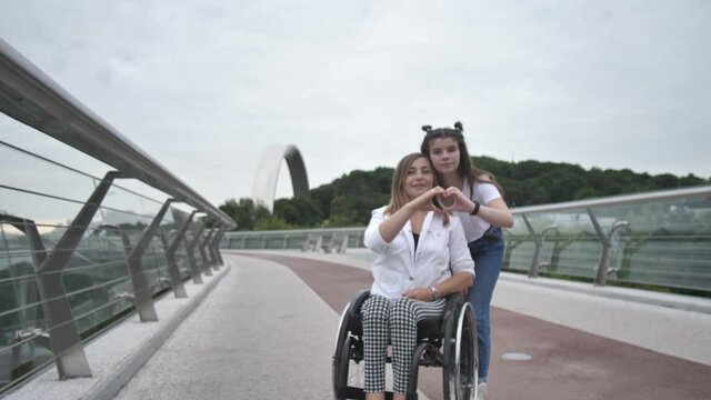 Smiling adult handicapped woman on wheelchair and her pretty teen daughter making heart shape with their fingers during walk on city footbridge. Love, togetherness, positive lifestyle with disability