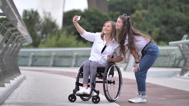 Smiling teen daughter and disabled mother on wheelchair posing for photo on footbridge. Happy women using smartphone while taking selfie pictures on background of city park nature