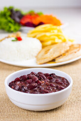 Black beans on white plate on raw cotton dish towel with a full plate with rice chicken and fries on background. feijoada