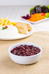 Black beans on white plate on raw cotton dish towel with a full plate with rice chicken and fries on background. feijoada
