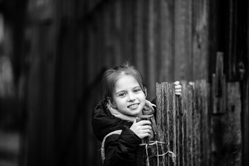 Portrait of a little girl behind a wooden fence in a village. Black and white photography.