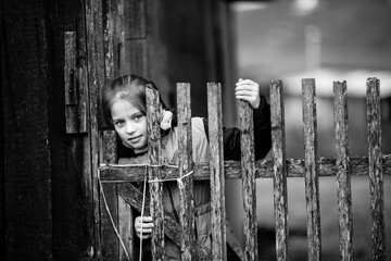 Portrait of a cute girl behind a wooden fence in the courtyard of a village house. Black and white photography.
