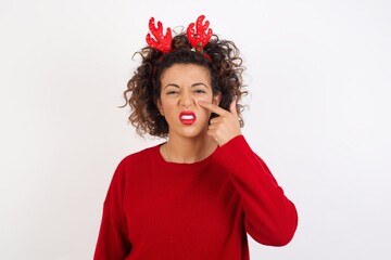 Young arab woman with curly hair wearing christmas headband standing on white background pointing unhappy to pimple on forehead, ugly infection of blackhead. Acne and skin problem