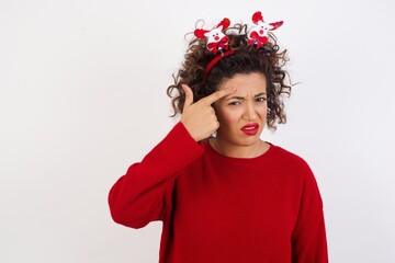 Young arab woman with curly hair wearing christmas headband standing on white background pointing unhappy at pimple on forehead, blackhead  infection. Skincare concept.