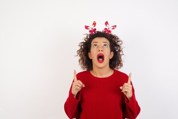 Young arab woman with curly hair wearing christmas headband standing on white background amazed and surprised looking up and pointing with fingers and raised arms.