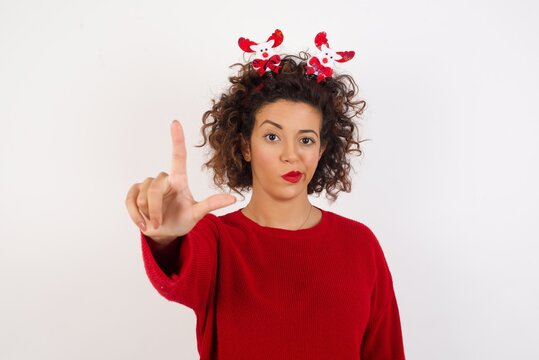 Young arab woman with curly hair wearing christmas headband standing on white background making fun of people with fingers on forehead doing loser gesture mocking and insulting.