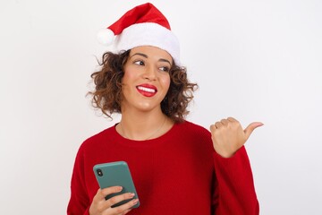 Young arab woman with curly hair wearing christmas headband on white background using and texting with smartphone  pointing and showing with thumb up to the side with happy face smiling