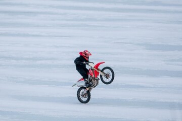 St. John's, Newfoundland/Canada - January 2020: A young man drives a red dirt bike over a lake that is frozen with thick ice. The boy is doing wheelies on the motorcycle. He is wearing a red helmet.