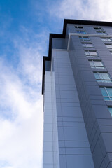 The corner top of a tall modern building with multiple coloured grey metal panels. There's a blue sky with white clouds in the background. There are small closed windows in the dark grey panels.  