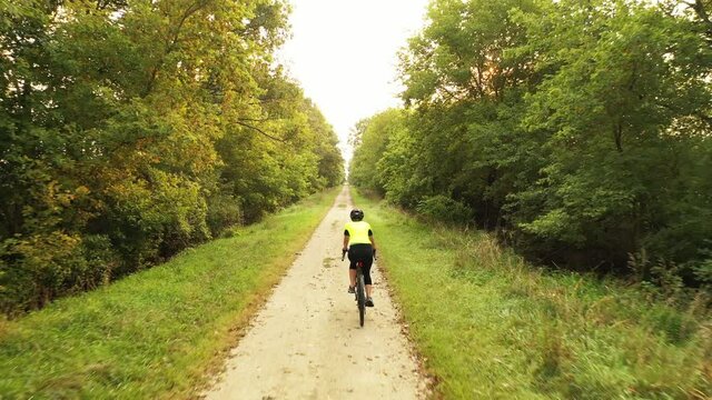 Cyclist is riding rail trail in american countryside. Riding bike on a rural road. Touring, adventure, travel concept. Morning sun is shining through trees, sun flare. Drone following, tracking shot 