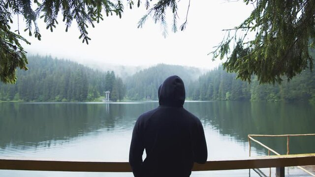 Tourist guy in black hoodie wearing and standing on pier of cinematic lake on cold spring morning. Inspiration shot. Beautiful pristine nature. In the mountains.