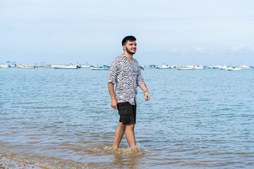 Hombre joven Adolescente con mascarilla caminando feliz  por la orilla del mar en la playa 