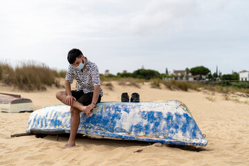 Adolescente con mascarilla limpiándose las pies de arena en la playa sentado en una barca