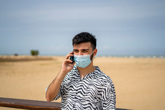 Hombre joven con mascarilla llamando por telefono movil en paseo maritimo junto a la playa y mar