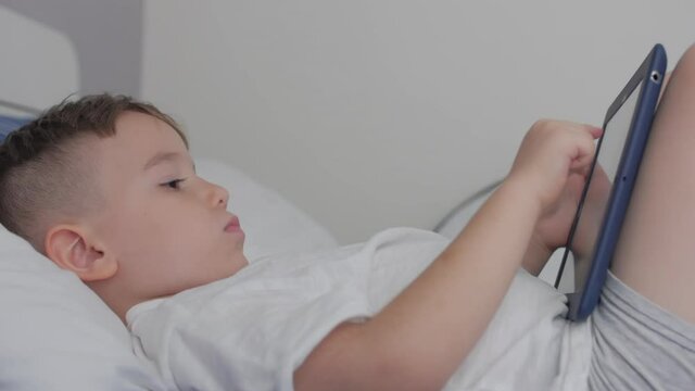 Cute little boy preschool child using a phone or digital tablet technology device looks telephone,plays in the phone sitting rest lying on sofa alone.Children tech addiction concept play game at home.