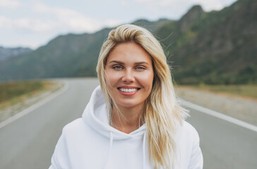Beautiful smiling blonde young woman traveler in white hoodie on road, trip to the mountains, Altai