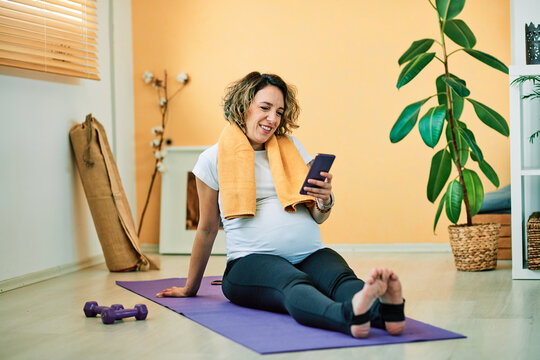 Attractive middle aged pregnant yogi woman sitting on the mat and using smart phone.