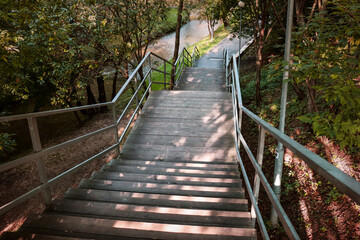 Fototapeta na wymiar Ladder in park with metal handrails. Wooden steps going down among trees, river in background. Climbing stairs in fresh air, early autumn. Sunlight, shadows from foliage