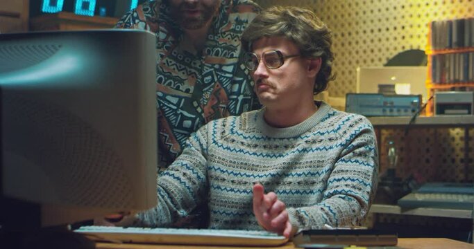 Caucasian retro stylish man in glasses and with moustache sitting at PC monitor and talking with colleague or friend. Vintage hackers of 80's. Nerd hacker from 70's working on computer with co-worker.