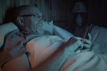 Closeup of senior man yawns while using digital tablet in bed before go to sleep. His wife sleep at background