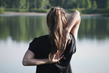 Rear view of young flexible woman does a hand lock behind her back. Close-up of woman practising yoga on lake shore