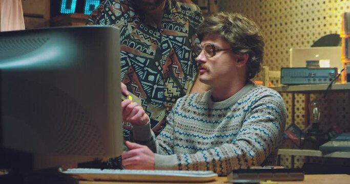 Caucasian man nerd in glasses and with mustaches from 70's sitting at PC monitor and discussing diskette with friend. Males hackers at computer quarreling and talking at home in retro interior.